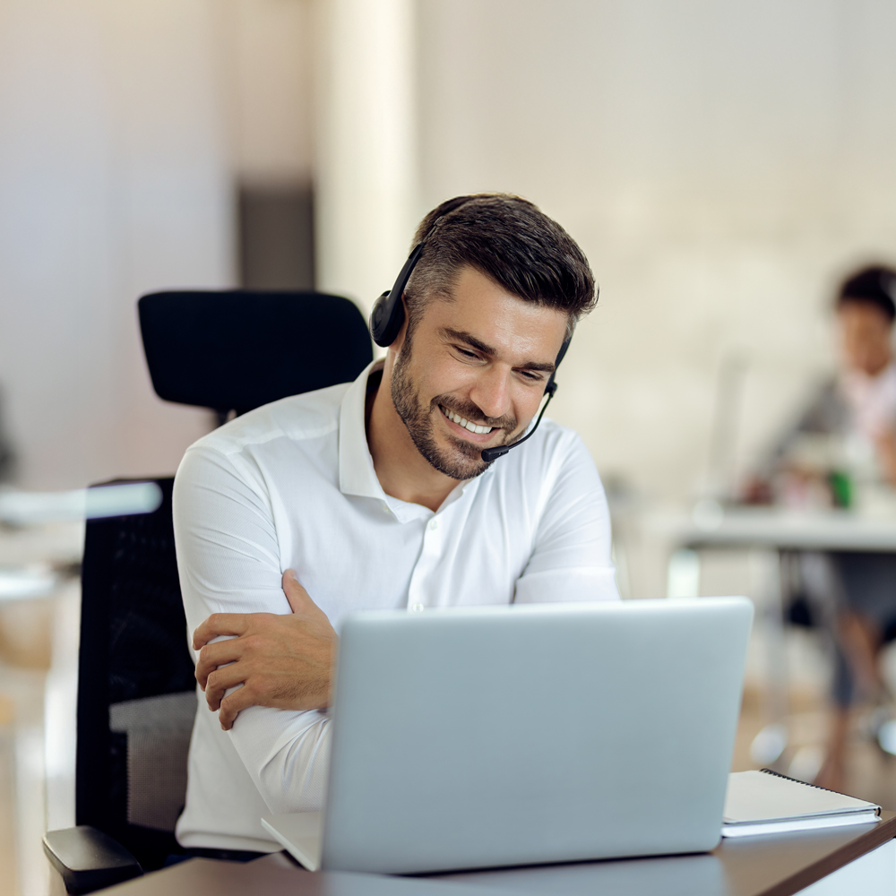A man smiling while working on a laptop in a modern office.
